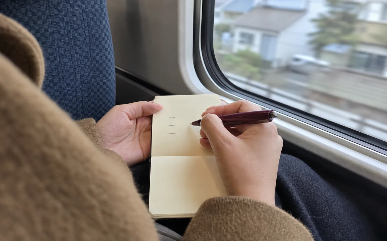 電車の窓辺で小さなノートに3項目を書く手元